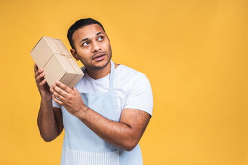 Fototapeta premium Portrait of positive attractive african american black indian cook man in apron looking at camera isolated over yellow background. Holding package.