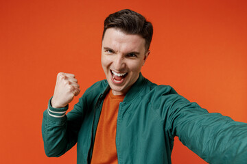 Close up young brunet man 20s wears red t-shirt green jacket doing selfie shot pov on mobile phone clench fists say yes isolated on plain orange background studio portrait. People emotions concept