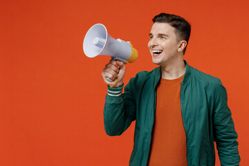 Excited charismatic smiling cheerful young brunet man 20s wears red t-shirt green jacket hold scream in megaphone announces discounts sale Hurry up isolated on plain orange background studio portrait