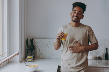 Young sated african american man in casual clothes drink orange juice put hand on belly eat muesli...