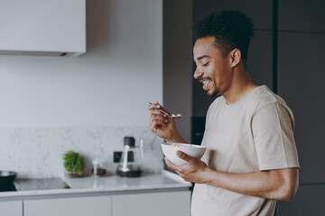 Side view young happy smiling african american man in casual clothes eat breakfast muesli cereals...