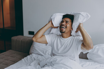 Angry sad tired young african american man in nightwear sitting in bed cover ears with pillow from...