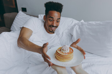 Young fun african american man in nightwear lying in bed receive take girlfriend breakfast pancakes...