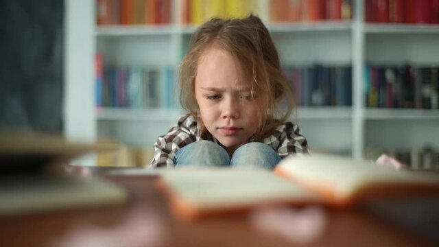 Front view of exhausted upset primary child school girl sitting alone hugging knees in front of desk with difficult homework. Upset angry schoolgirl put head on knee being stressed with homework