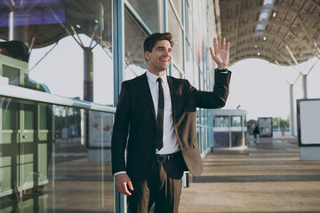 Bottom view friendly young traveler brunet businessman man 20s wearing black classic tie suit standing outside at international airport terminal look aside waving hand Air flight business trip concept