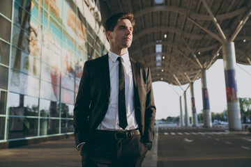 Bottom view minded young traveler brunet businessman man 20s in black classic tie suit standing outside at international airport terminal look aside. People air flight business trip lifestyle concept.