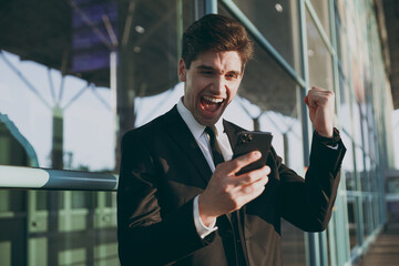 Young traveler businessman man in black suit stand outside at international airport terminal use mobile cell phone book taxi order hotel do winner gesture clench fist Air flight business trip concept.