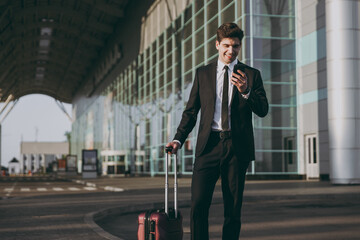 Bottom side view traveler businessman young man in black suit stand outside at international airport terminal with suitcase talk on mobile phone book taxi order hotel Air flight business trip concept