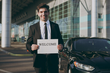 Bottom view young smiling friendly traveler businessman man 20s in black suit stand outside at international airport terminal hold card sign with welcome title text Air flight business trip concept