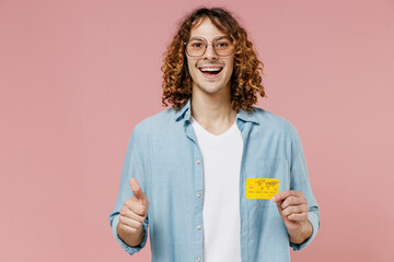 Young smiling satisfied happy man with long curly hair in blue shirt white t-shirt glasses hold in hand credit bank card show thumb up isolated on pastel plain pink color background studio portrait