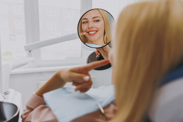 Young smiling satisfied fun happy calm woman 20s reflected in mirror sitting at dental office chair indoor light cabinet look at teeth toothy smile whitening result. Healthcare caries enamel treatment