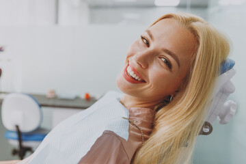 Close up young smiling happy calm blonde woman covered by napkin sit at dental office chair indoor light modern cabinet waiting stomatologist for oral procedure. Healthcare caries enamel treatment. - obrazy, fototapety, plakaty