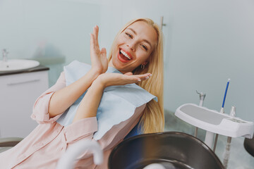 Young amazed smiling happy woman 20s covered by napkin spread hands sit at dental office chair...