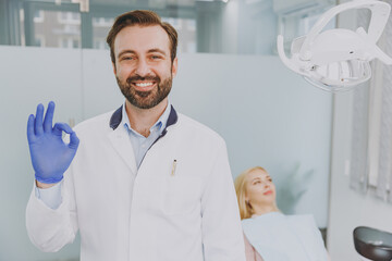 Obraz premium Young confident smiling happy male man doctor show ok okay gesture stand near patient woman sitting at dentist office chair indoor light cabinet near stomatologist Healthcare oral enamel treatment.