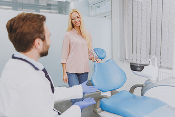 Young blonde caucasian satisfied happy calm cheerful woman 20s enter dental office indoor light cabinet, male european doctor stomatologist say sit down., Healthcare oral enamel aesthetic treatment