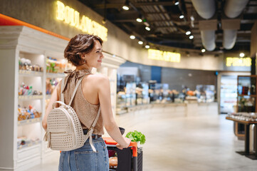 Young smiling happy european woman 20s wear casual clothes shopping at supermaket store with grocery cart look aside produce inside hypermarket. People lifestyle purchasing gastronomy food concept