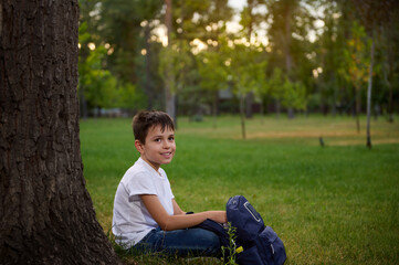 Adorable handsome child school boy resting sitting on green grass at the park, enjoying recreation after school. Portrait of happy clever elementary aged kid boy student with backpack. Back to school