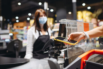 Cropped close up woman hand in casual clothes mask shopping at supermaket with grocery cart put mobile phone to modern bank payment terminal process acquire payment near cashier checkout inside store