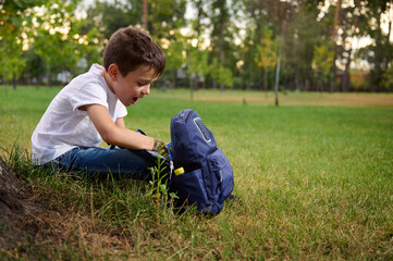 Naklejka na ściany i meble Elementary aged school boy in casual denim resting on green grass after his first day at school. Adorable child with school bag during recreation on beautiful sunny day. Back to school concept