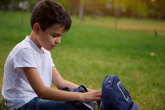 Close-up portrait of an adorable schoolboy with an open backpack sitting in the park during a break after school and looking for work books and supplies in a school bag. Back to school concept - Powered by Adobe