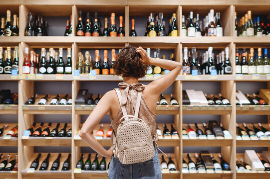 Back view puzzled minded young woman in casual clothes shopping at supermaket grocery store buy choosing wine alcohol bottle scratch head inside hypermarket. People purchasing gastronomy food concept.