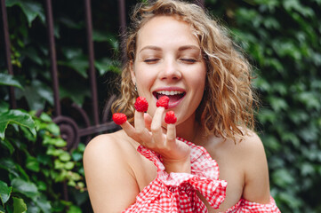 Close up young wistful happy satisfied woman in pink dress put girl put raspberries on fingers eat...
