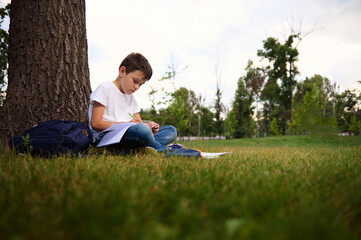 Obraz premium Serene child schoolboy concentrating on doing homework, sitting on the city park green grass, enjoying fresh air during his recreation after first day at school. Back to school concept. September.