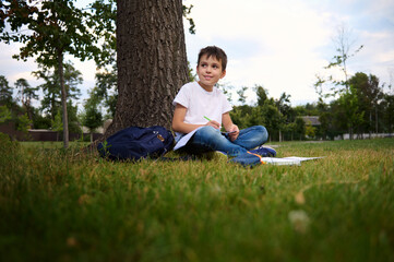 Naklejka na ściany i meble Serene handsome school boy doing homework, sitting on city park green grass, looking to the side, enjoying fresh air during his recreation after first day at school. Back to school concept. September.