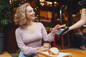 Young fun woman in casual clothes at cafe buy breakfast sit at table hold wireless modern bank...