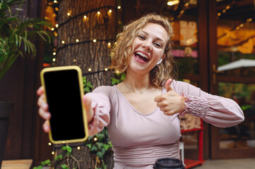 Young smiling woman in casual clothes sit alone at table in cafe shop listen music in earphones...