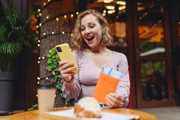 Young amazed happy tourist woman in casual clothes sitting alone at table in cafe use mobile cell phone hold passport boarding tickets eat breakfast relax in restaurant before traveling abroad indoor.