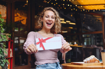 Young happy smiling woman in casual clothes sitting alone at table in coffee shop cafe eat breakfast hold gift certificate coupon voucher card for store relaxing in restaurant during free time indoor - obrazy, fototapety, plakaty