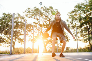 Young serious sunlit sporty concentrated sportsman man 20s in sports clothes hood covering with palms hands hold in hand ball play at basketball game playground court Outdoor courtyard sport concept