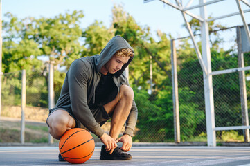 Fototapeta premium Young sporty caucasian sportsman man 20s wearing grey sportswear hood looking aside training sit laces up sneakers shoes with ball at basketball game playground court Outdoor courtyard sports concept