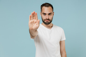 Young sad strict man 20s in casual white t-shirt looking camera showing stop gesture with palm refusing isolated on plain pastel light blue color background studio portrait. People lifestyle concept