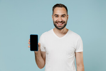 Young smiling happy fun man in blank print design white t-shirt hold in hand mobile cell phone with blank screen workspace area isolated on plain pastel light blue color background studio portrait