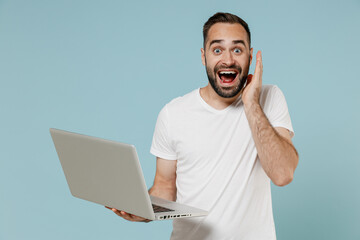 Young shocked surprised excited happy fun man 20s wearing casual basic white t-shirt hold use work on laptop pc computer hold face isolated on plain pastel light blue color background studio portrait