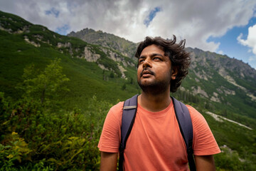 Obraz premium Portrait of a focused adventurer Indian tourist man looking up towards the way forward with bag pack against mountains during hike. Summer adventure and sports concept