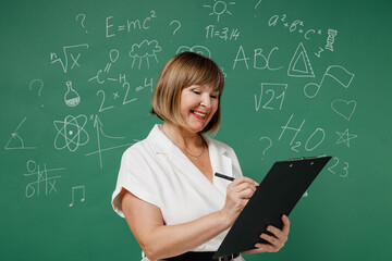 Happy teacher mature elderly lady woman 55 wear white shirt hold write in clipboard with papers document isolated on green wall chalk blackboard background studio. Education in high school concept.