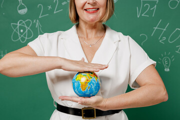 Close up cropped fun teacher mature elderly lady woman 55 wear white shirt hold in palm Earth world globe isolated on green wall chalk blackboard background studio. Education in high school concept