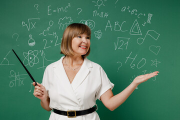 Overjoyed teacher mature elderly lady woman 55 wear white shirt look aside demonstrate with pointer palm isolated on green wall chalk blackboard background studio. Education in high school concept.