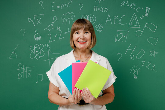 Smiling intelligent fun teacher mature elderly lady woman 55 wear white shirt hold in hand colour papers isolated on green wall chalk blackboard background studio. Education in high school concept