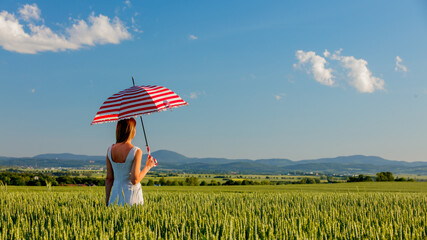 young girl in hat with umbrella on green wheat field and mountains on background