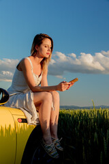 young girl in sunglasses with smartphone is sitting on a car on green wheat field in spring time