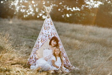 Family portrait of lovely caring mother hugging her adorable daughter, while sitting in front of decorated wigwam tent at sunset in summer wild field © sofiko14