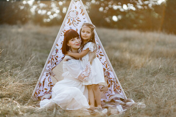 Close up outdoor shot of likable lovely mother and little daughter, wearing family look white dress and feathers in hair, posing together in the field. Teepee wigwam on the background © sofiko14