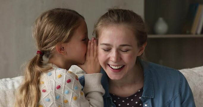 Little girl share secrets with mom, close up. Cheerful curious woman listens to cute adorable 9s daughter telling confidential information on ear. Trusting relations, closeness, communication concept
