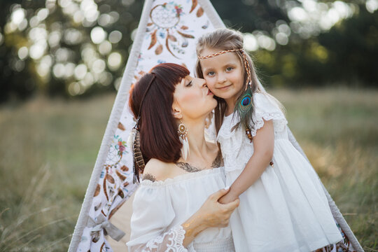 Family shot in native american indin style. Happy young mother cuddles and kisses her daughter in the summer on field, sitting near boho tee pee tent. Mom and daughter wearing white dress and feathers