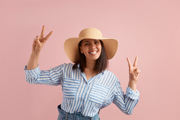 Obraz na płótnie Canvas Smiling positive woman with dark hair shows peace sign with both hands, feels excited to go on vacation, wears straw hat, striped shirt, jeans with belt, on pink background. Summer emotions concept.