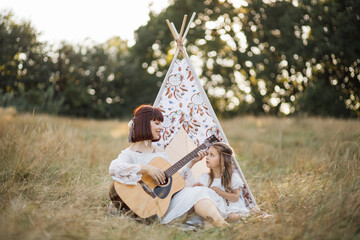 Pretty young mother and daughter playing guitar together, sitting next to teepee tent outdoors in...